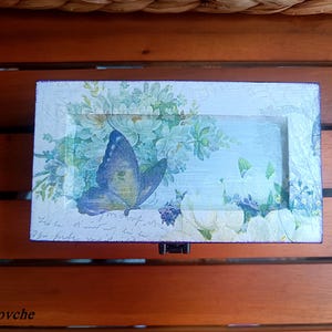 Puede incluir: Una peque&ntilde;a caja de madera con una mariposa azul y un dise&ntilde;o floral. La caja est&aacute; pintada de blanco con un acabado desgastado y tiene una mariposa azul con alas blancas y amarillas. La caja est&aacute; decorada con un dise&ntilde;o floral en tonos azules y verdes.