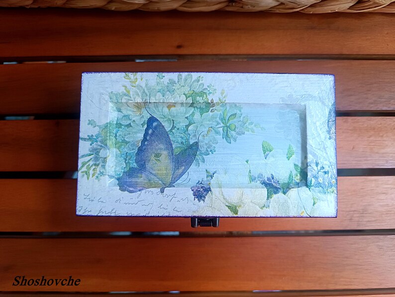 Puede incluir: Una peque&ntilde;a caja de madera con una mariposa azul y un dise&ntilde;o floral. La caja est&aacute; pintada de blanco con un acabado desgastado y tiene una mariposa azul con alas blancas y amarillas. La caja est&aacute; decorada con un dise&ntilde;o floral en tonos azules y verdes.