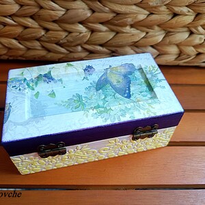 Puede incluir: Una peque&ntilde;a caja de madera con un acabado pintado en morado y blanco. La caja tiene un dise&ntilde;o floral con una mariposa en la tapa. La caja tiene dos bisagras de lat&oacute;n.