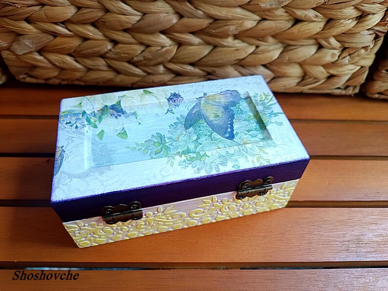 Puede incluir: Una peque&ntilde;a caja de madera con un acabado pintado en morado y blanco. La caja tiene un dise&ntilde;o floral con una mariposa en la tapa. La caja tiene dos bisagras de lat&oacute;n.