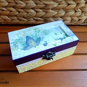 Puede incluir: Una peque&ntilde;a caja de madera con un dise&ntilde;o pintado en morado y blanco. La tapa presenta un patr&oacute;n floral con una mariposa azul. La caja tiene un cierre de metal.