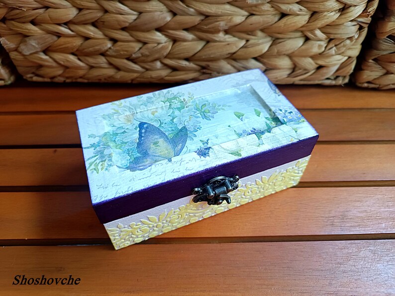 Puede incluir: Una peque&ntilde;a caja de madera con un dise&ntilde;o pintado en morado y blanco. La tapa presenta un patr&oacute;n floral con una mariposa azul. La caja tiene un cierre de metal.
