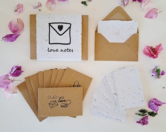 May include: A stationery set featuring handmade paper and brown envelopes. The set includes notecards, envelopes, and a larger card with the words "love notes" and a heart-shaped design. Pink flowers are scattered around the set.