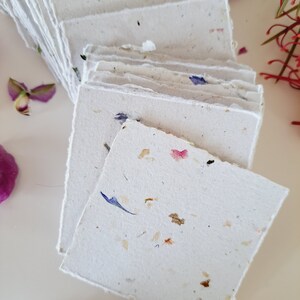 May include: A stack of handmade paper cards with embedded flower petals. The cards are white with a rough texture and have a variety of colors of flower petals embedded in them.