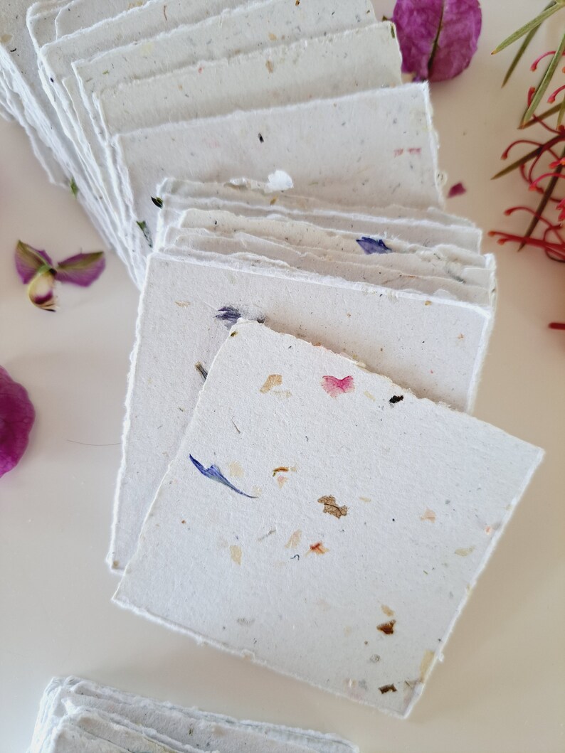 May include: A stack of handmade paper cards with embedded flower petals. The cards are white with a rough texture and have a variety of colors of flower petals embedded in them.