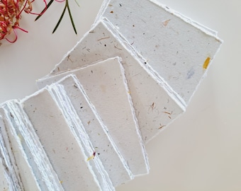 May include: A stack of handmade, square paper sheets with deckled edges. The off-white paper is embedded with small flecks of dried plant material in shades of brown, yellow, and purple. The paper is arranged in a fan shape, with a few sheets visible.