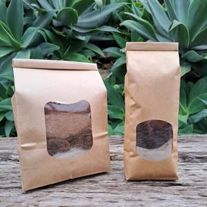 May include: Two brown paper bags with a clear window showing brown and white wool inside. The bags are sitting on a wooden surface.