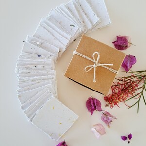 May include: A stack of white handmade paper with flecks of dried flowers, arranged in a semi-circle. A small brown box with a twine bow sits in the center of the circle. Pink and purple dried flowers are scattered around the box and paper.