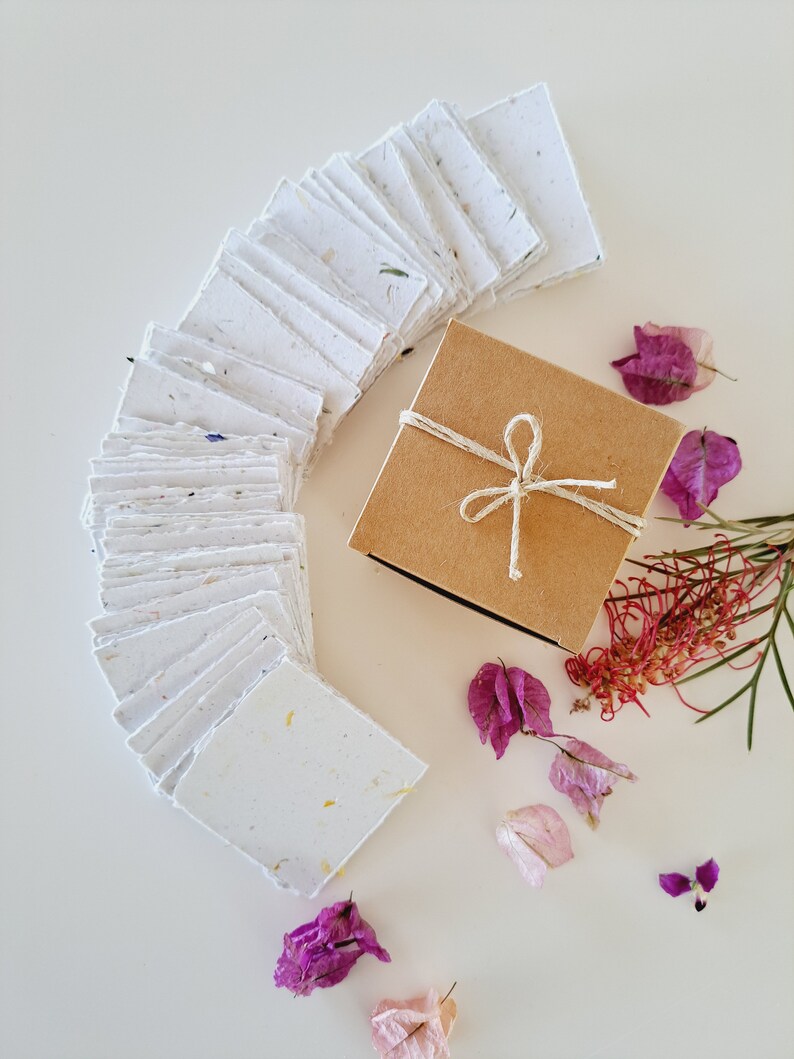 May include: A stack of white handmade paper with flecks of dried flowers, arranged in a semi-circle. A small brown box with a twine bow sits in the center of the circle. Pink and purple dried flowers are scattered around the box and paper.