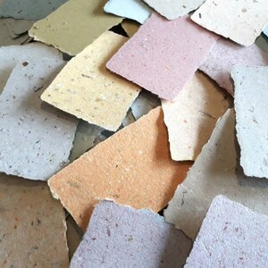May include: A pile of handmade paper sheets in various colors, including beige, light brown, light blue, and pink. The paper has a textured surface and is made from recycled materials.