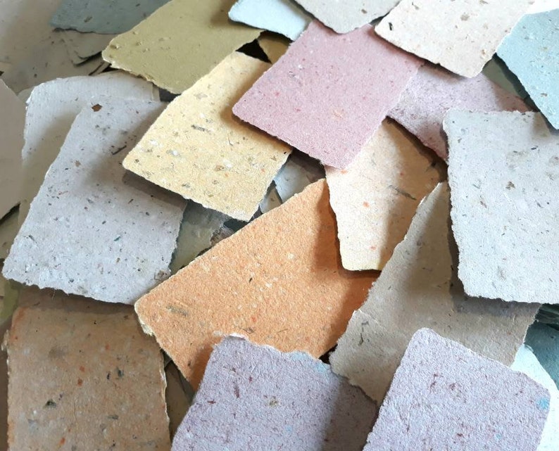 May include: A pile of handmade paper sheets in various colors, including beige, light brown, light blue, and pink. The paper has a textured surface and is made from recycled materials.