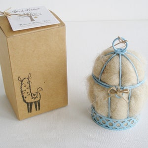 May include: A light blue metal bird cage filled with natural wool fibers. The cage is sitting on a white surface. The cage is packaged in a brown cardboard box with a llama illustration and the text "Bird Nester Natural Wool Fiber".