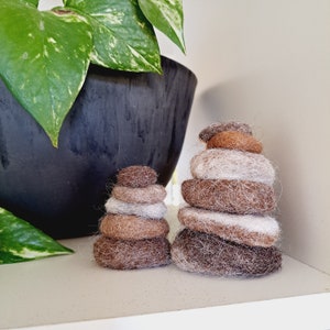 May include: Two stacks of brown and white felt stones, resembling a zen garden, on a white shelf. A green leafy plant is in a black pot behind the stones.