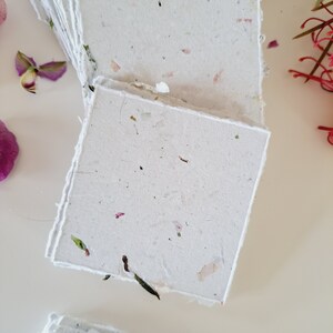 May include: A stack of handmade paper cards with a white background and flecks of dried flowers and leaves. The cards have a rough, textured surface.