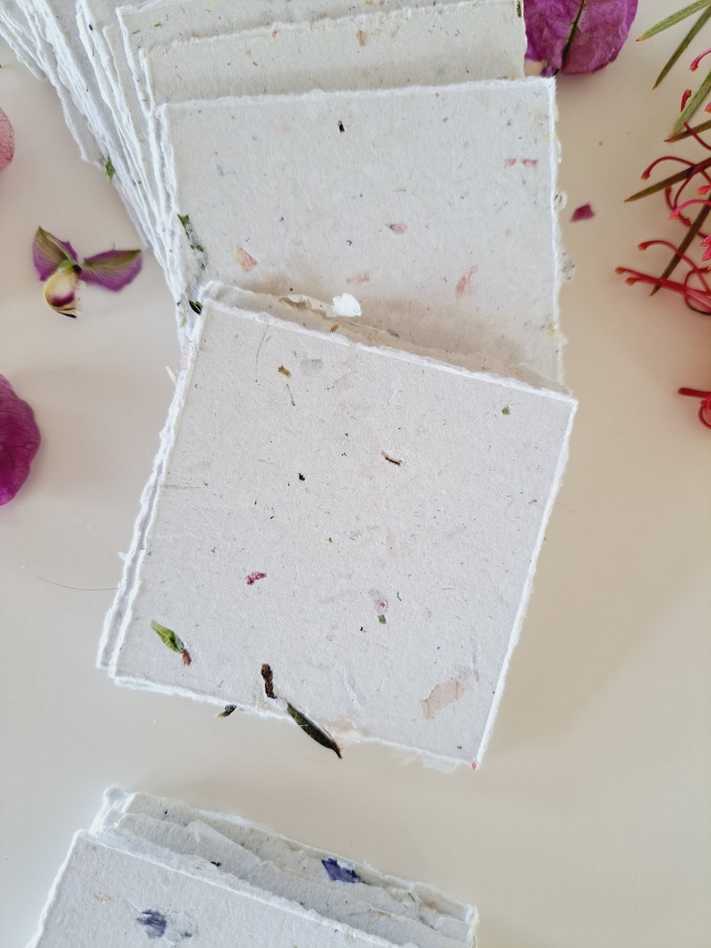 May include: A stack of handmade paper cards with a white background and flecks of dried flowers and leaves. The cards have a rough, textured surface.