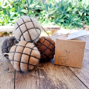 May include: Four brown, beige and white felted wool balls with wire cages. The balls are packaged in a brown box with a llama illustration and the text "The Alpaca Tree".