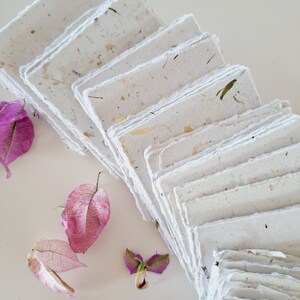 May include: A stack of handmade paper sheets with a textured, speckled surface. The paper is white with flecks of brown, green, and yellow. The paper is arranged in a fan shape, with the edges of the sheets overlapping. The paper is surrounded by pink flower petals.