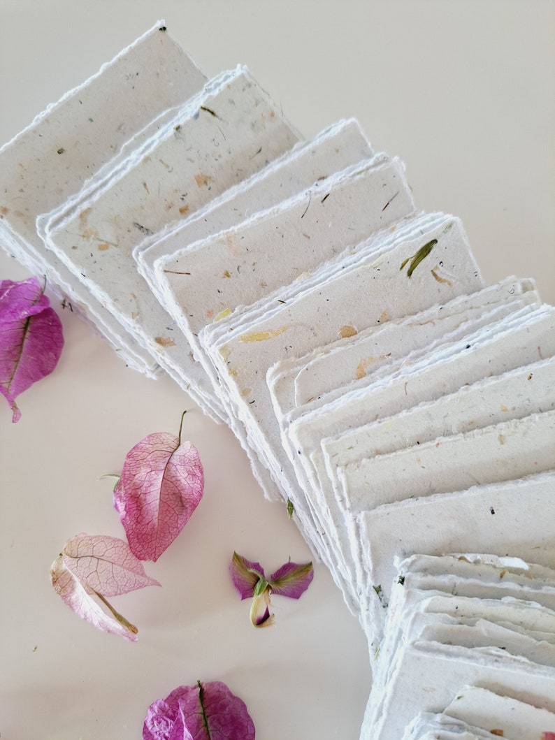 May include: A stack of handmade paper sheets with a textured, speckled surface. The paper is white with flecks of brown, green, and yellow. The paper is arranged in a fan shape, with the edges of the sheets overlapping. The paper is surrounded by pink flower petals.