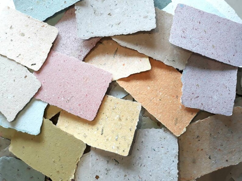 May include: A collection of handmade paper scraps in various colors, including light blue, beige, pink, yellow, and brown. The paper has a textured, speckled appearance, suggesting it is made from recycled materials.