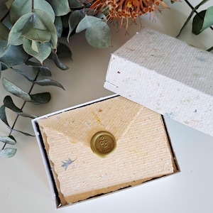 May include: A rectangular white box containing a stack of light tan, textured handmade paper envelopes with deckled edges. A single envelope is sealed with a round, metallic gold wax seal featuring a small envelope icon with hearts.