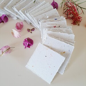 May include: A stack of white handmade paper cards with a speckled texture. The cards are arranged in a semi-circle with the top card facing the viewer. The cards are surrounded by pink and purple flowers.