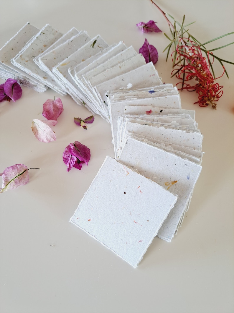 May include: A stack of white handmade paper cards with a speckled texture. The cards are arranged in a semi-circle with the top card facing the viewer. The cards are surrounded by pink and purple flowers.