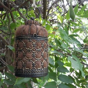 May include: A brown and gold metal birdhouse with a decorative design hangs from a tree branch. The birdhouse is filled with brown nesting material.