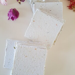 May include: A stack of handmade paper sheets with a textured surface and flecks of natural materials. The paper is white with a subtle off-white hue.