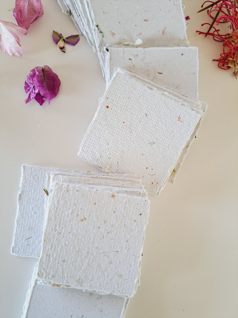 May include: A stack of handmade paper sheets with a textured surface and flecks of natural materials. The paper is white with a subtle off-white hue.