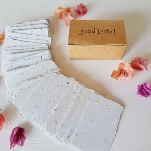 May include: A stack of handmade, rectangular white paper cards with flecks of colour, arranged in a curved pattern. A small, brown box with the words "good Luck!" is next to the cards. Dried pink and orange flowers are scattered around the cards and box.