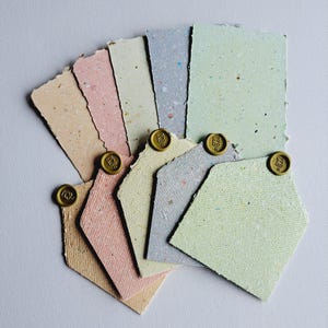 May include: A collection of rectangular handmade paper sheets in peach, pink, cream, grey, and light green, each featuring deckled edges and flecks of fibre. Four of the sheets are folded into envelopes and sealed with small, round, gold wax seals.