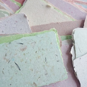 Pu&ograve; includere: Un mucchio di fogli di carta fatti a mano in vari colori pastello, tra cui rosa, verde, blu e bianco. La carta ha un aspetto testurizzato e macchiato, il che suggerisce che sia fatta di materiali riciclati.
