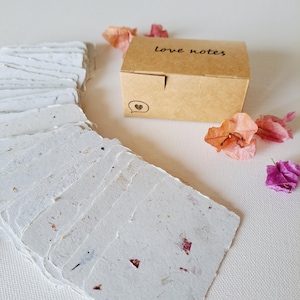 May include: A collection of handmade white paper cards with a textured finish and floral inclusions. A small brown box, labelled "love notes" with a heart icon, is positioned nearby. Dried pink and orange flowers are scattered around the cards and box.