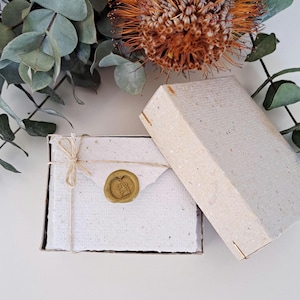 May include: A rectangular box made of textured off-white paper containing a matching envelope tied with thin twine. The envelope is sealed with a gold-colored wax seal featuring a small heart and envelope icon. The box lid sits beside it.