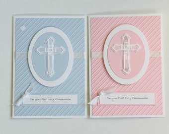 May include: Two handmade cards with a white cross design in an oval frame. The card on the left is light blue with white diagonal stripes. The card on the right is pink with white diagonal stripes. Both cards have the text "On your First Holy Communion" printed on them.