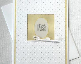 May include: A white greeting card with a yellow border and a textured, dotted pattern. The center features a yellow gingham square with an oval illustration of purple flowers. A white ribbon bow is tied at the bottom.