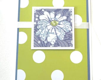 May include: A handmade greeting card with a daisy design. The card features a lime green background with white polka dots, a blue border, and a square floral design in the center. A white ribbon is wrapped around the card.