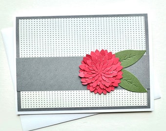 May include: A handmade greeting card with a red paper flower and green leaves. The card has a gray band and a white background with a dotted pattern. An envelope is included.