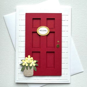 May include: A handmade greeting card featuring a red door with a gold "WELCOME" sign. The card has a white brick-like background and a basket of yellow flowers. A white envelope is behind the card.