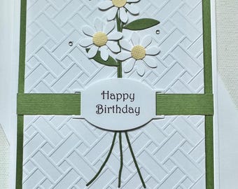 May include: A white Happy Birthday greeting card with a textured background and a green border. The card features a bouquet of white daisies with yellow centers and green leaves. The words "Happy Birthday" are printed on a white oval.