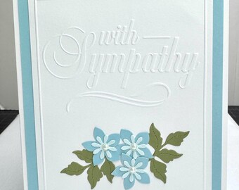 May include: A sympathy card with a white background and a light blue border. The card features embossed text that reads "with sympathy" and a floral arrangement of blue flowers with pearl centers and green leaves.