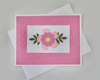 May include: A handmade greeting card with a pink embossed background featuring a floral design. The card has a white border and a white rectangular insert with a pink flower, green leaves, and small rhinestones. A white envelope is included.