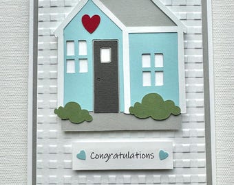 May include: A white card with a gray border and a blue house with a red heart on the front. The house has a gray door and two windows. The card says "Congratulations" in blue text with two blue hearts.