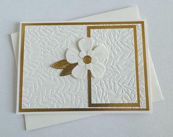 May include: A white card with a gold border and a white paper flower with a gold center. The card has a white embossed pattern of leaves.