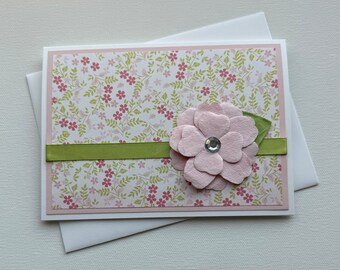 May include: A handmade greeting card with a pink floral design and a green ribbon. The card features a pink paper flower with a rhinestone center.