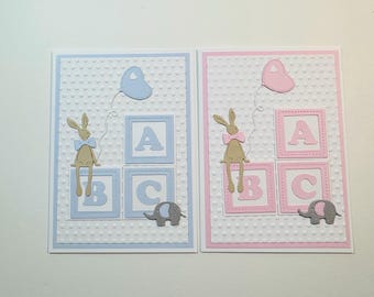 May include: Two greeting cards with a white background and a light blue and pink border. Each card features a bunny rabbit holding a balloon, sitting on top of alphabet blocks that spell ABC. The cards are perfect for a baby shower or new baby.