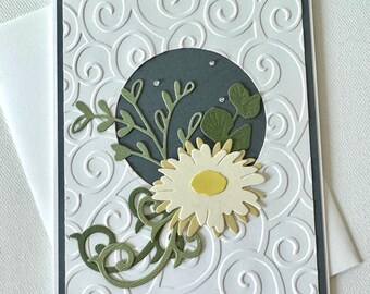 May include: A handmade greeting card with a white embossed background featuring swirling patterns. A dark gray circle holds a floral arrangement with a cream-colored daisy, green leaves, and a yellow center. The card is framed in dark gray.
