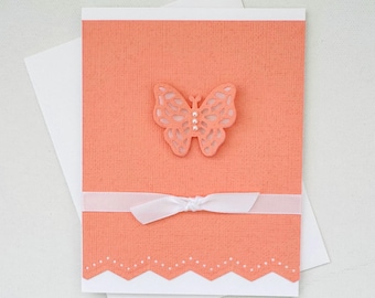 May include: A coral-colored greeting card with a textured surface, featuring a decorative butterfly and a white ribbon tied in a bow. The card has a scalloped edge and is paired with a white envelope, perfect for special occasions.