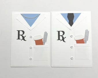 May include: Two white greeting cards designed to resemble lab coats. Each card features a blue collar, a pocket with a coffee cup and pen, and the Rx symbol. One card has a pearl necklace, the other a black tie.