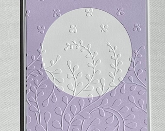 May include: A lavender card with a white circle in the center. The circle has a white embossed design of leaves and branches. The background of the card is also embossed with a swirling leaf design.